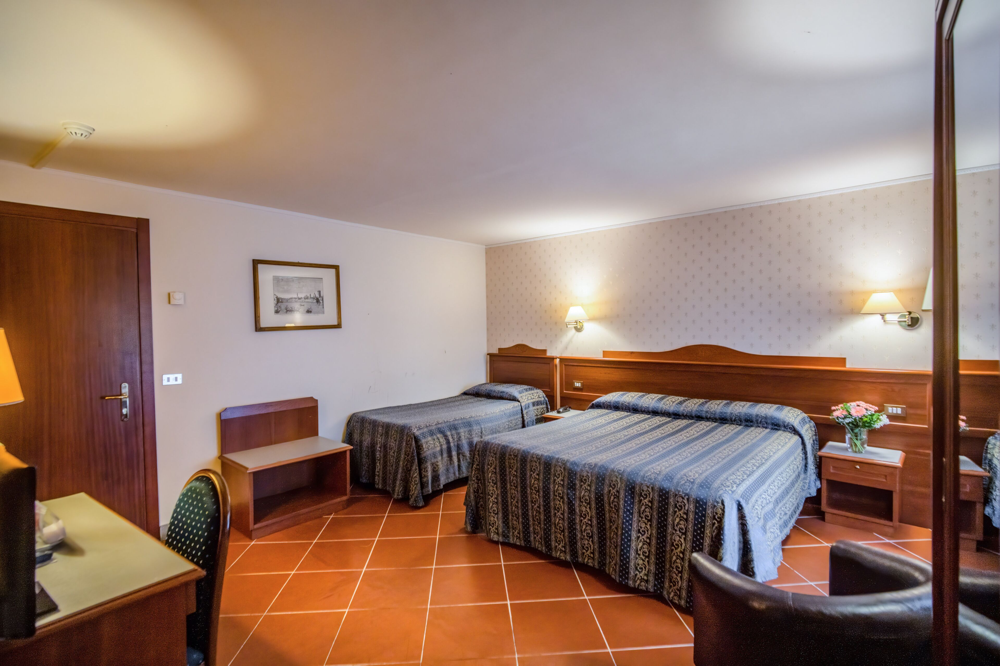 Une chambre d’hôtel avec deux lits simples, un bureau, une chaise, une petite table, une table de chevet et une lampe fixée au mur.