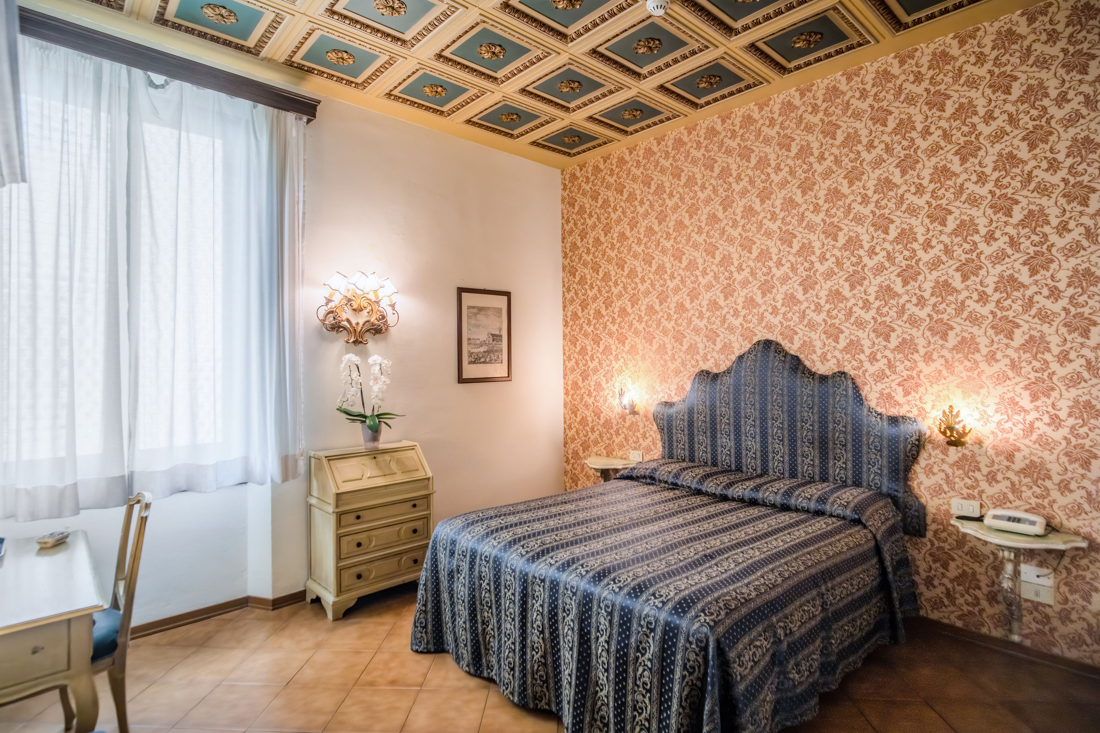 A bedroom with a patterned bedspread, a wooden nightstand, a wall-mounted lamp, and a window with sheer curtains.