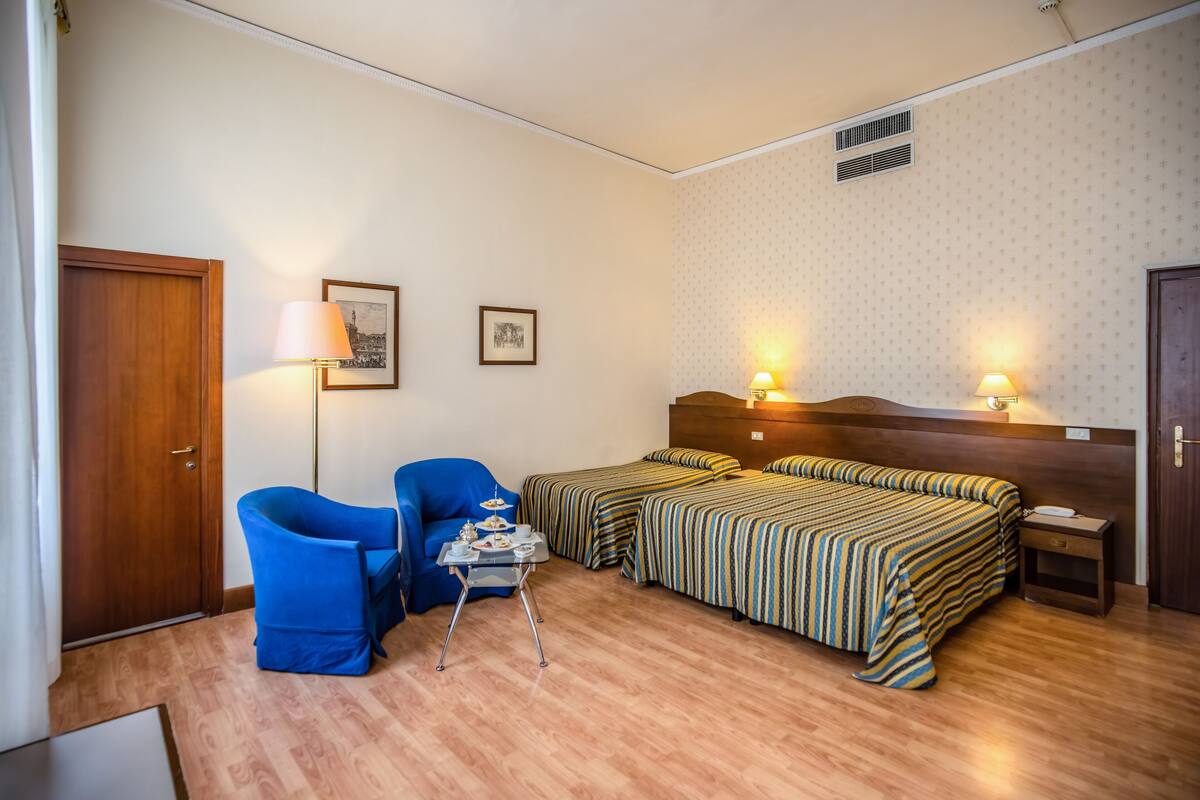 Hotellihuone, jossa on kaksi erillistä sänkyä, sininen nojatuoli, pieni pöytä lampun kanssa ja puinen ovi.