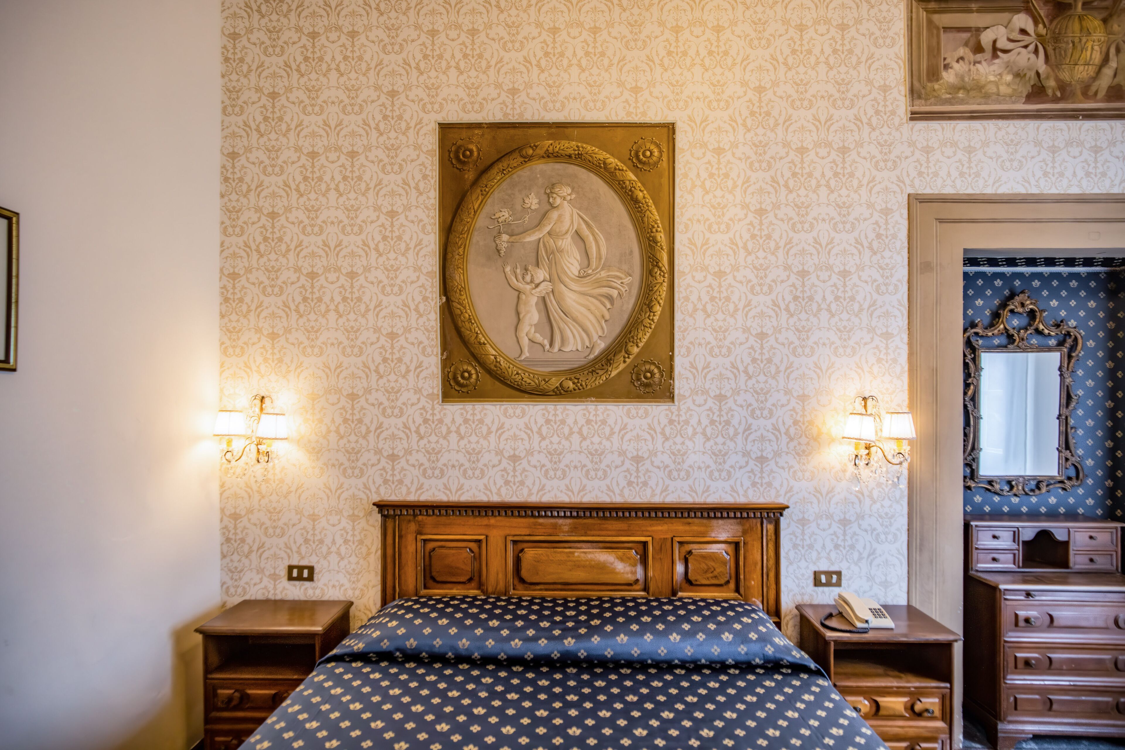 Quarto com cabeceira de madeira, cama com colcha estampada em azul e dourado, mesa de cabeceira com luminária, cômoda com espelho e um relevo decorativo fixado na parede.