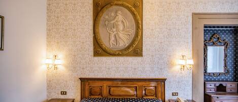 Quarto com cabeceira de madeira, cama com colcha estampada em azul e dourado, mesa de cabeceira com luminária, cômoda com espelho e um relevo decorativo fixado na parede.
