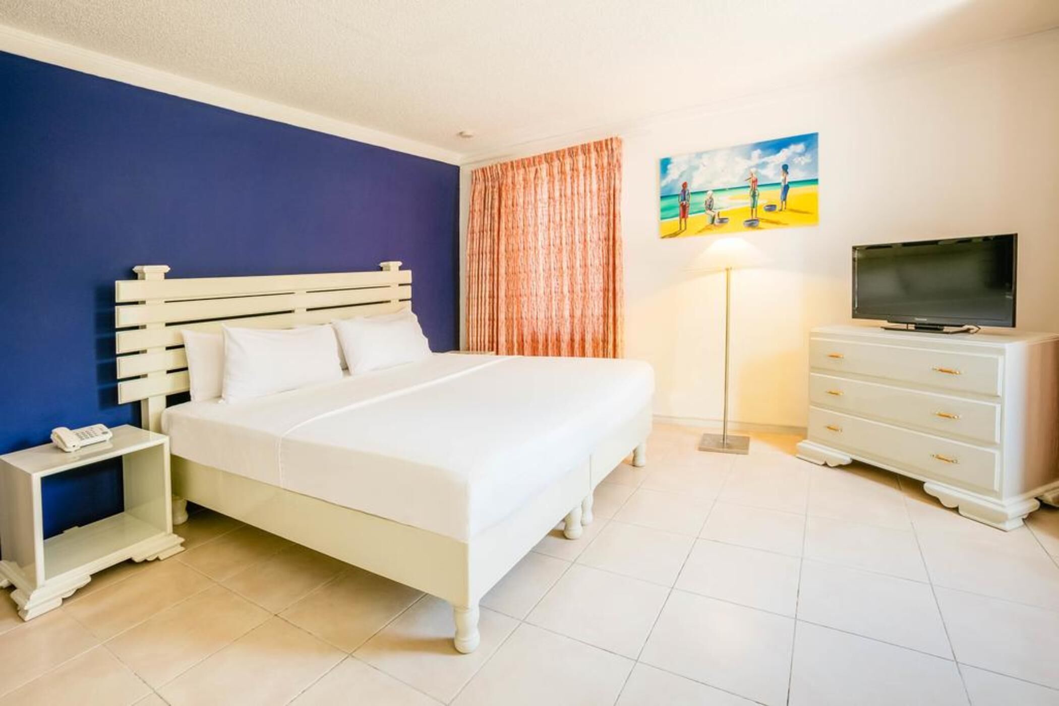 A hotel room with a bed, bedside table, lamp, TV, and a painting on the wall.