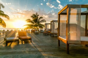 A wooden deck with lounge chairs and a glass-enclosed sleeping area, overlooking the ocean at sunset.