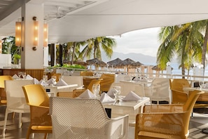 A restaurant with outdoor seating overlooking a beach, featuring wicker chairs and tables with white napkins and glassware. - Grand Decameron Montego Beach, A Trademark All Inc (Montego Bay)