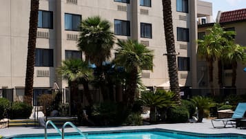 A swimming pool with a clear blue water surface, surrounded by palm trees and a multi-story building with balconies.