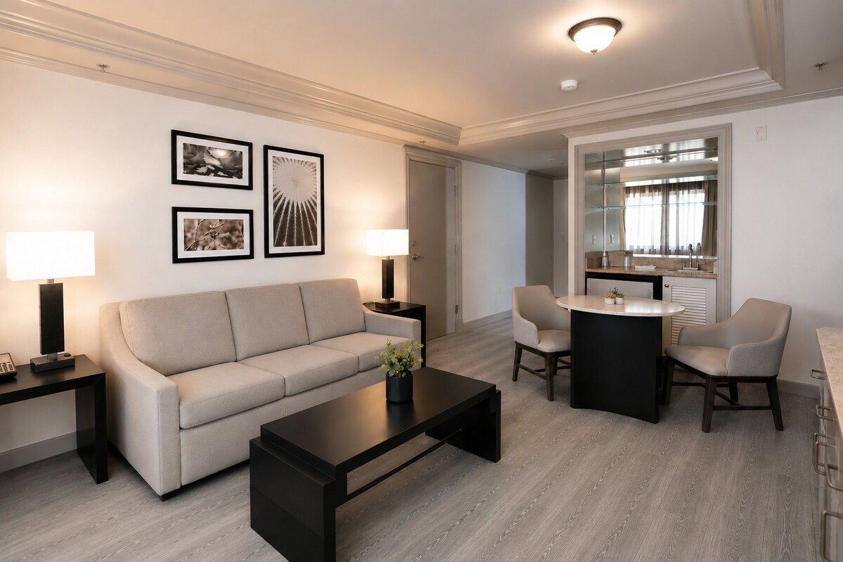 Een moderne woonkamer met een bank, salontafel en twee stoelen. Er hangen ingelijste foto's aan de muur, een lamp aan weerszijden van de bank, en een eethoek met een tafel en stoelen.