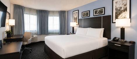 Ein Hotelzimmer mit einem großen Bett, einem Schreibtisch, einem Stuhl, einer Schreibtischlampe, einem Nachttisch mit Telefon und gerahmten Kunstwerken an der Wand.