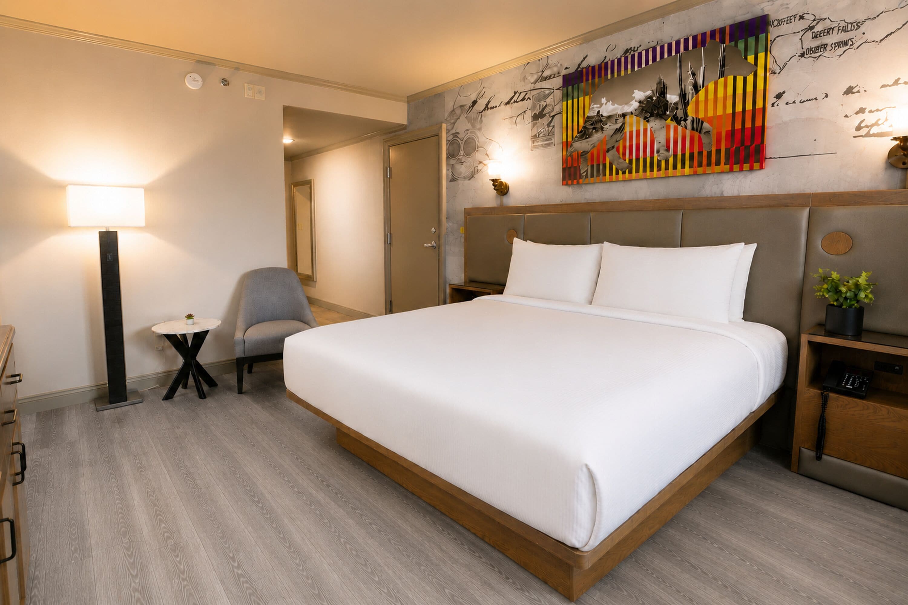 A hotel room with a large bed, a desk, a chair, a small table, a lamp, and a wall with artwork.