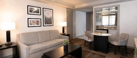 Habitación de hotel con sofá, mesa de centro, dos mesitas de noche con lámparas, zona de comedor con mesa redonda y sillas, y baño visible a través de una puerta de vidrio.