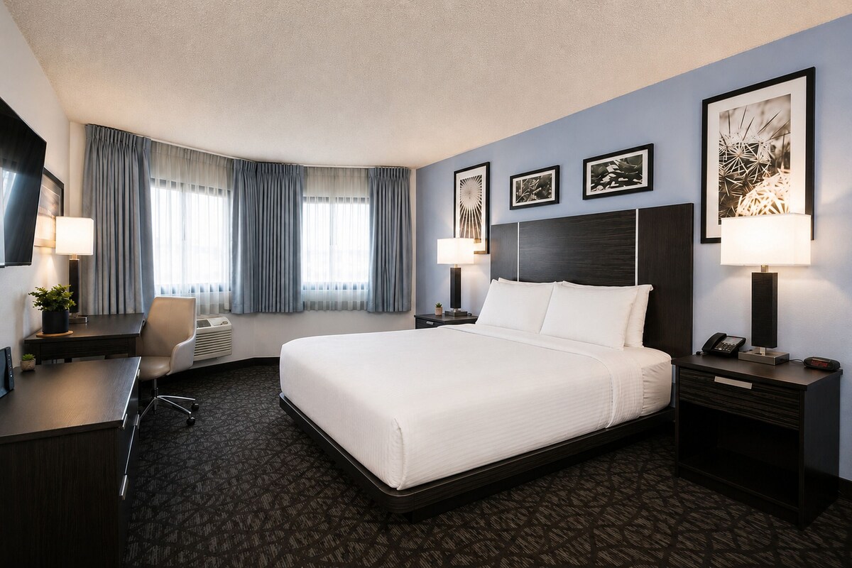 Een hotelkamer met een groot bed, een bureau met een stoel, een bureaulamp, een nachtkastje met een lamp en een aan de muur bevestigde televisie.
