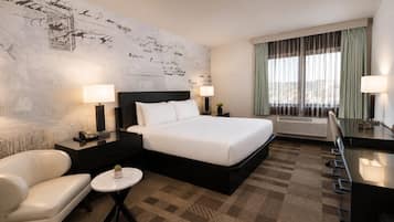 A hotel room with a large bed, two bedside tables with lamps, a small round table, a sofa, and a large window with curtains.