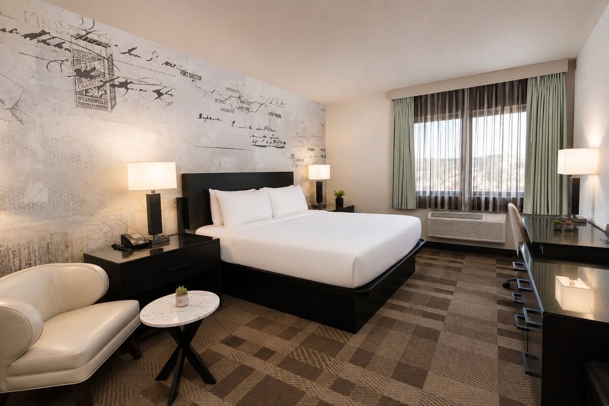 Een hotelkamer met een groot bed, twee nachtkastjes met lampen, een klein rond tafeltje, een bank en een groot raam met gordijnen.