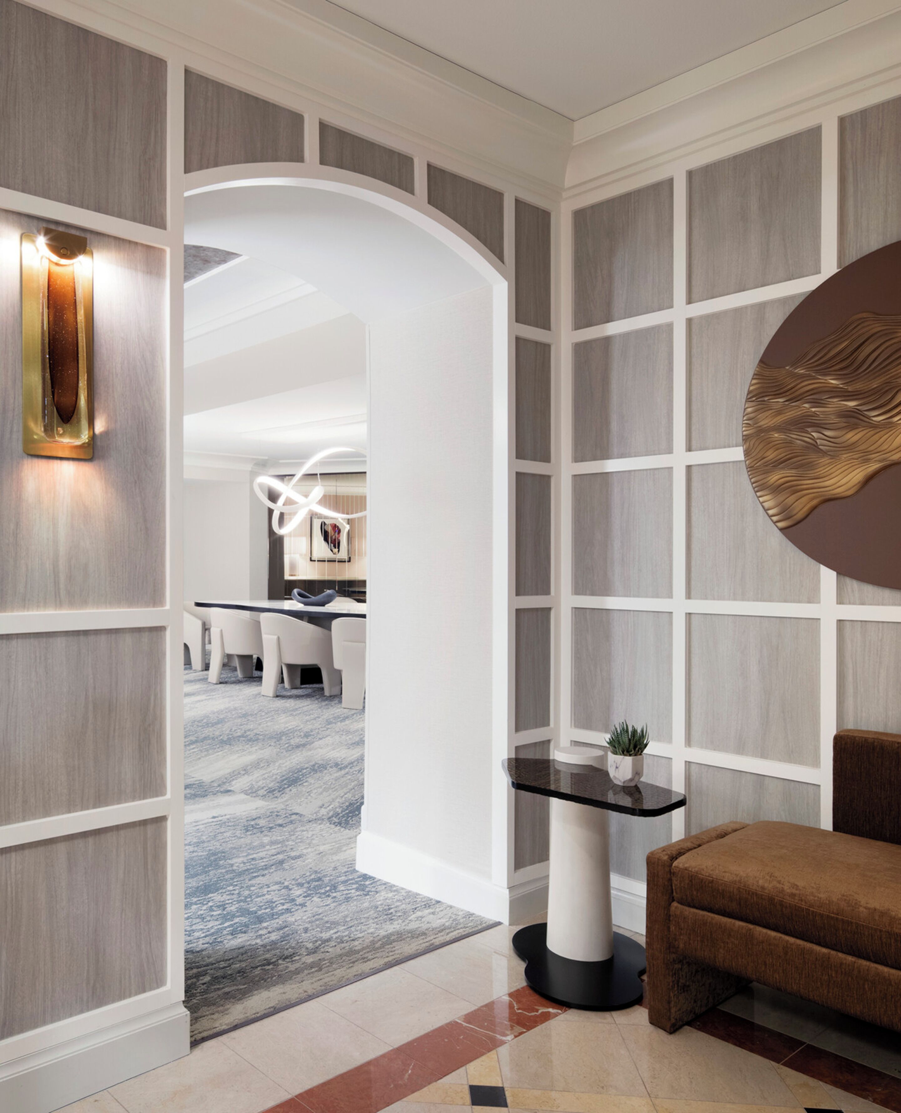 Un interior moderno con un pasillo que conduce a un comedor, que presenta una combinación de paneles de madera y paredes blancas, una pieza decorativa redonda en la pared y una pequeña mesa con una planta.