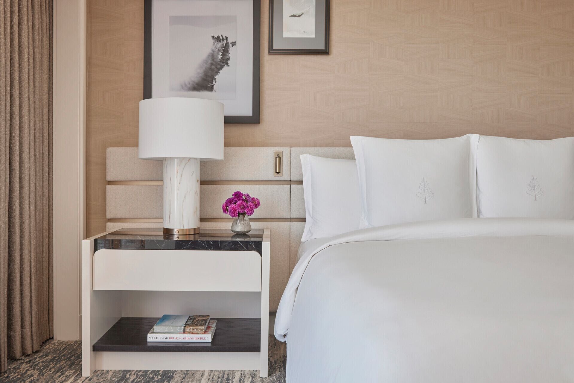 A hotel room with a bed, a nightstand with a lamp, a small table with a vase of flowers, and a framed picture on the wall.