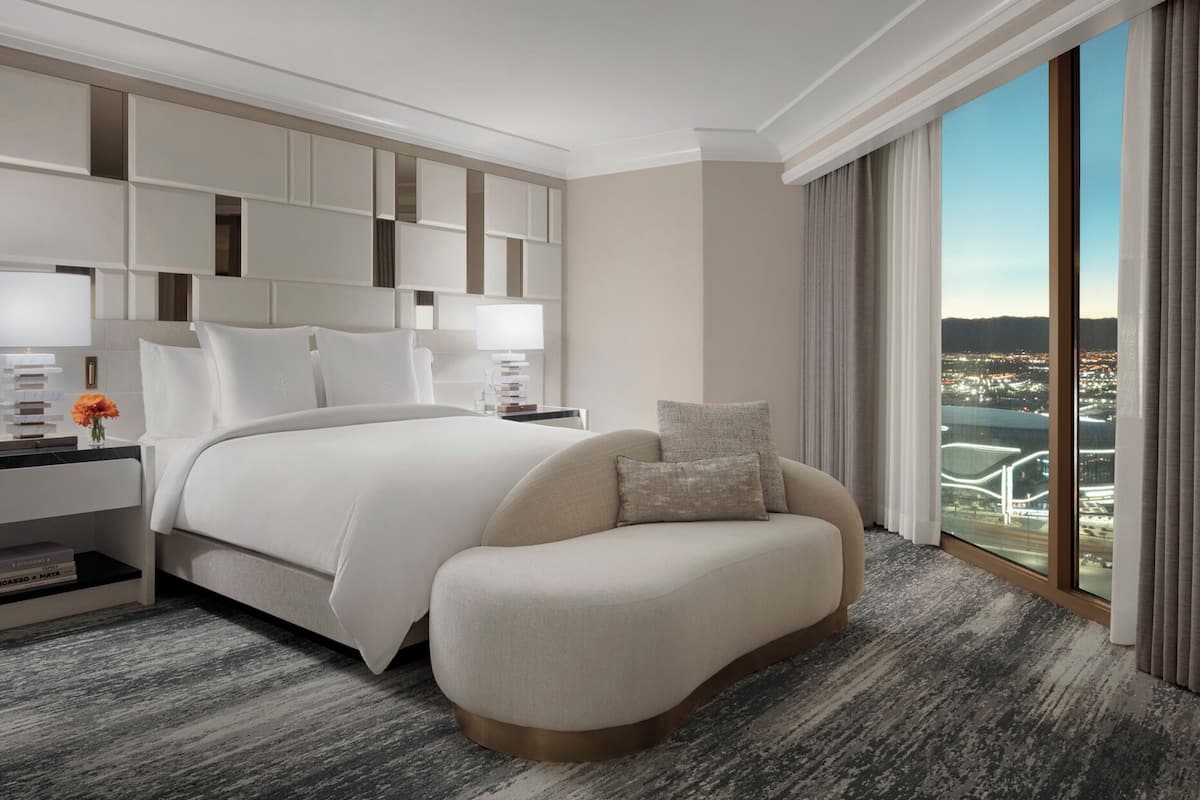 Una habitación de hotel moderna con una cama grande, un sofá, una mesita de noche y vistas a un paisaje urbano a través de amplias ventanas.