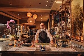 A bar area with a bartender, a marble counter, and shelves stocked with bottles and glasses.