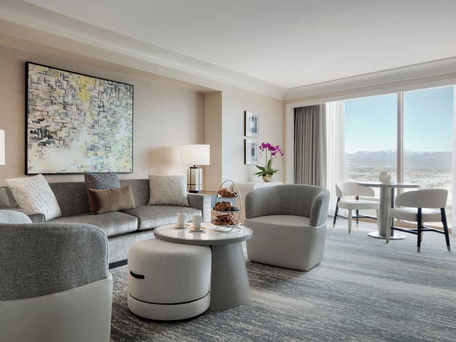 A modern hotel room with a grey sofa, armchairs, a round coffee table, and a dining area with a table and chairs.