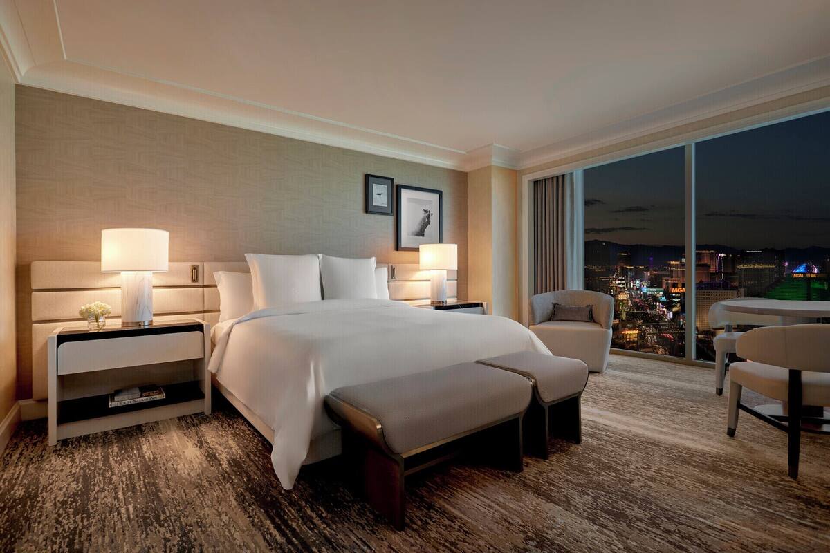 Una habitación de hotel con una cama grande, dos mesitas de noche con lámparas, un banco, una silla y un amplio ventanal con vistas a la ciudad.