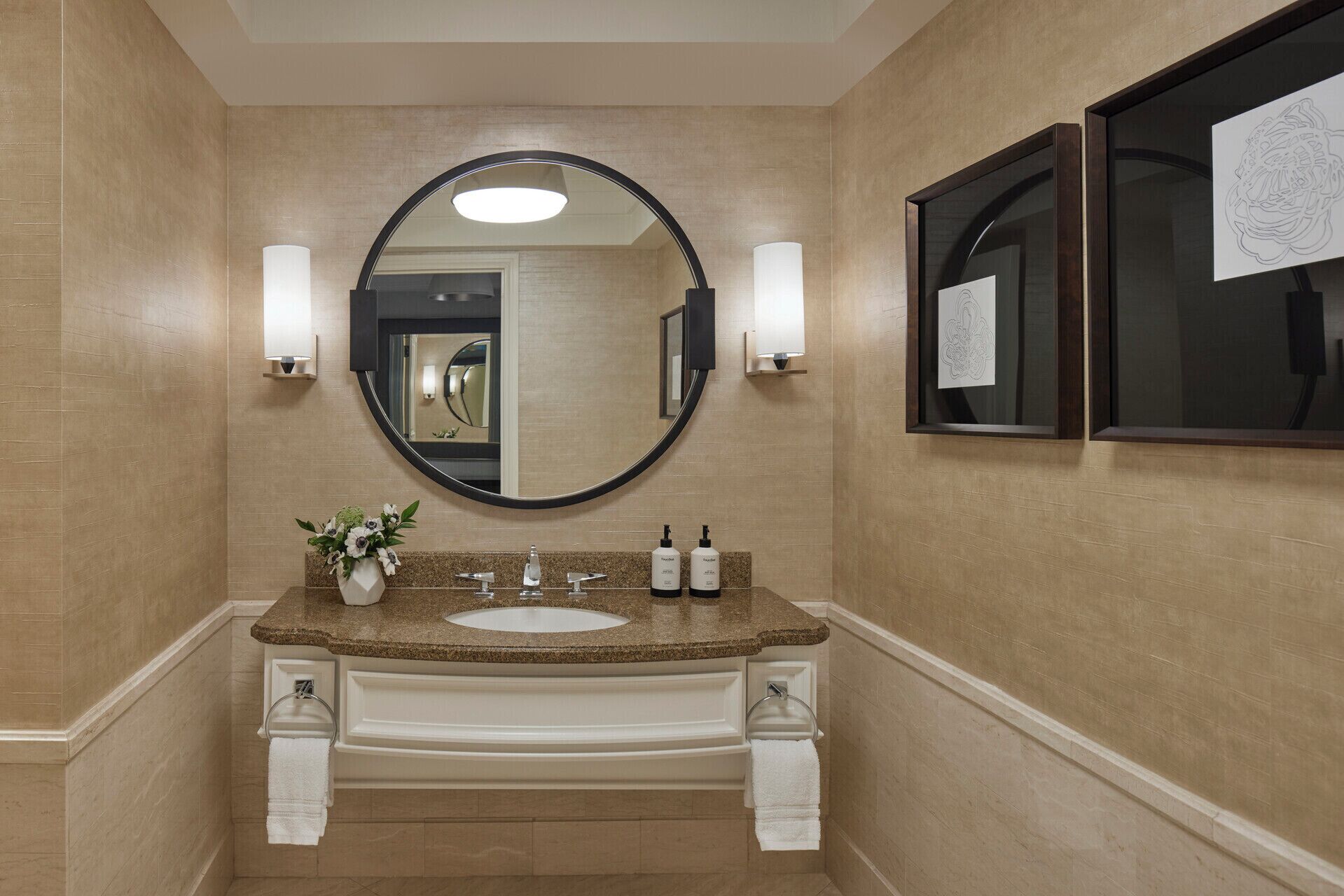 A bathroom with a round mirror, two wall-mounted lights, a granite countertop, a white sink, and a vase with flowers.