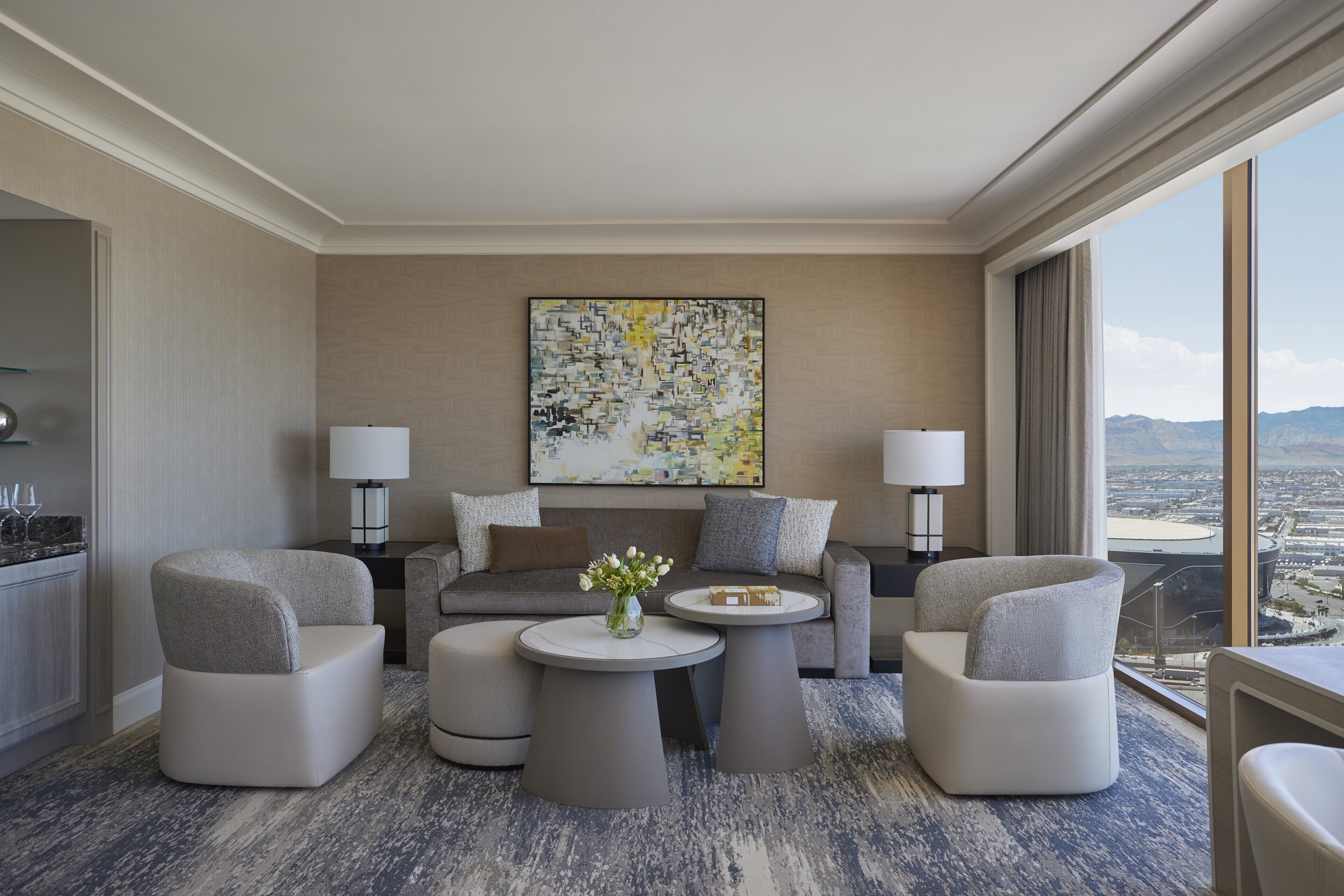 A modern hotel room with a large window offering a city view, a sofa set, two armchairs, a coffee table, and a painting on the wall.