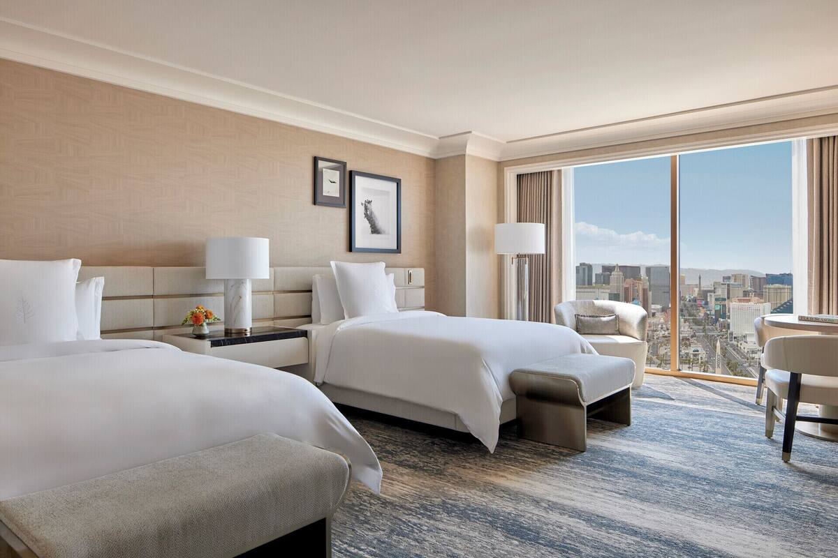 Una habitación de hotel con dos camas, un ventanal con vistas a la ciudad, un escritorio con lámpara y una silla.