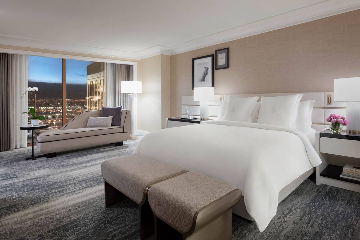 Habitación de hotel con una cama grande, un sofá, un banco, una mesita de noche y vistas a la ciudad.