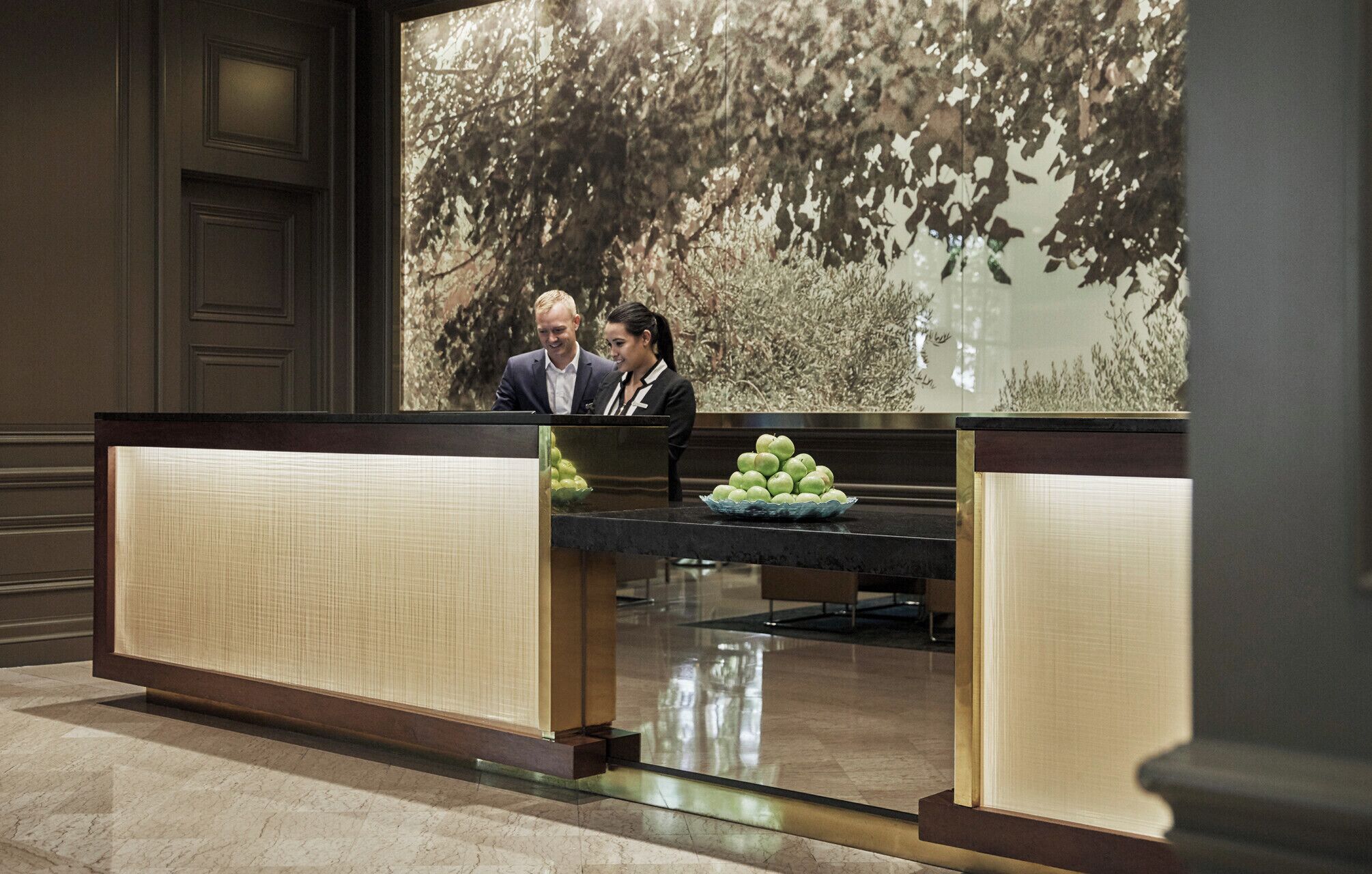 A reception desk with a large window displaying a forest scene. There are two people seated at the desk, and a bowl of green apples on the counter.
