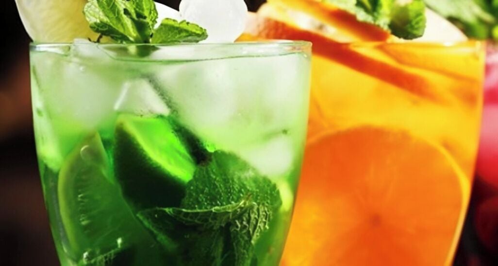 Un vaso de bebida verde con menta y lima, y una naranja en rodajas al fondo.