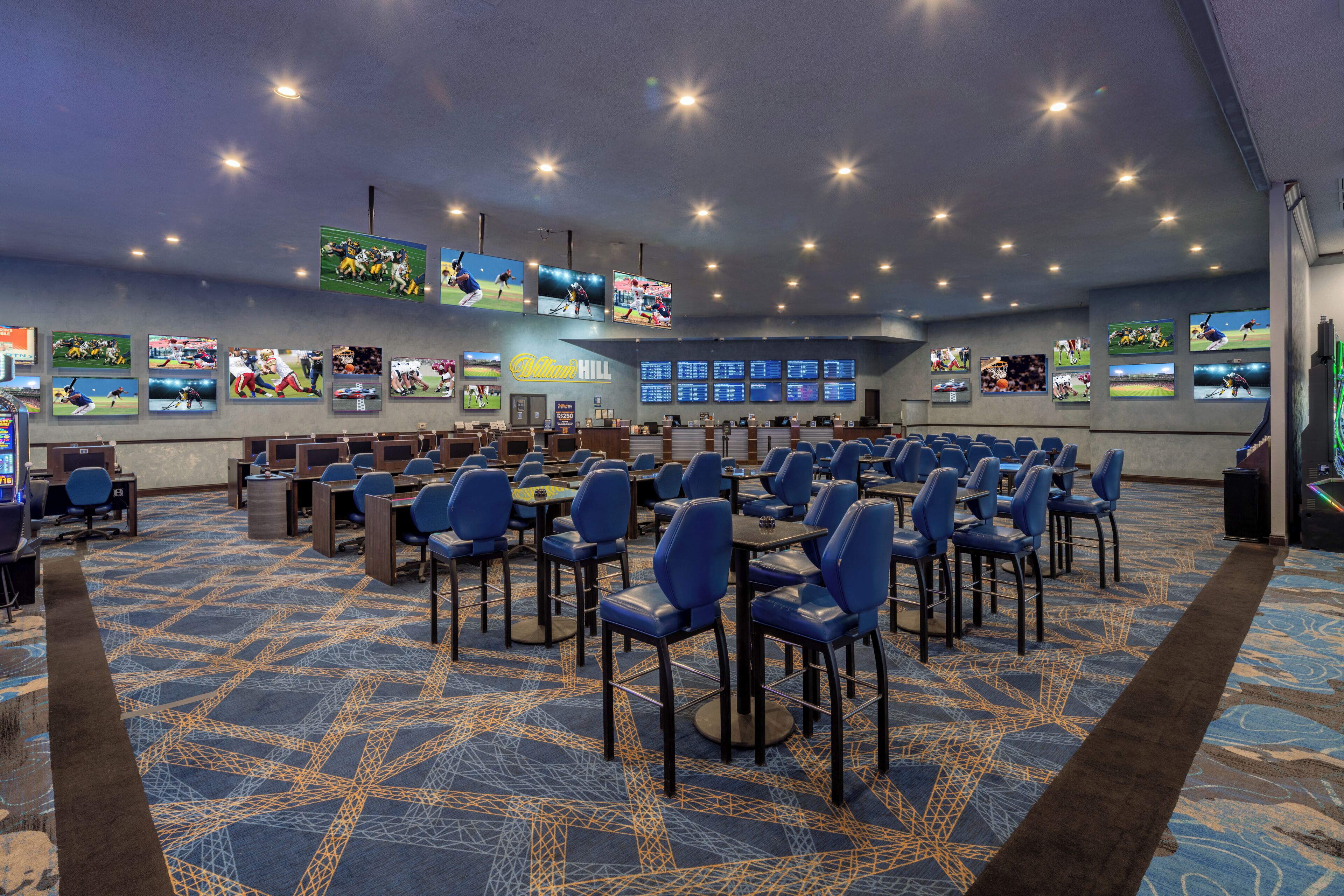 A conference room with multiple tables and chairs, digital screens displaying sports content, and a patterned carpet.