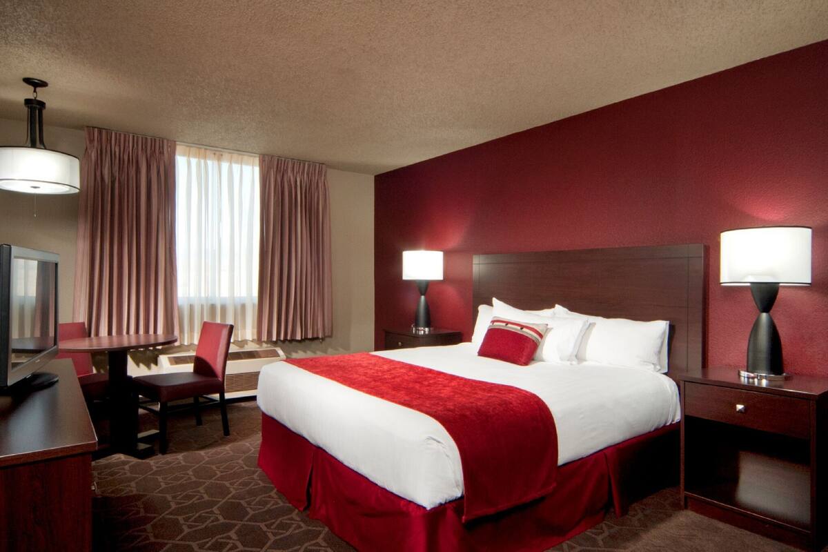 A hotel room with a large bed, two bedside tables with lamps, a desk with a chair, a television, and a window with curtains.