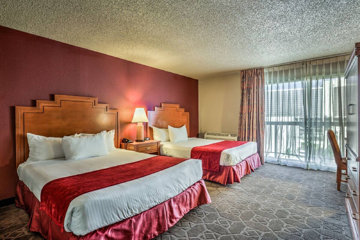 A hotel room with two beds, a nightstand with a lamp, a window with curtains, and a view of the city.