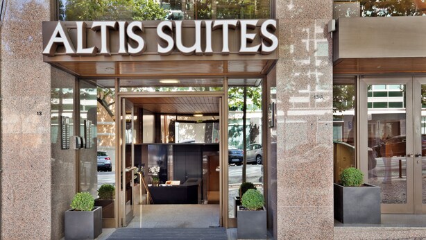 The exterior of a hotel named 'Altis Suites' with a modern entrance, glass doors, and potted plants.