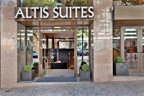 The exterior of a hotel named 'Altis Suites' with a modern entrance, glass doors, and potted plants.