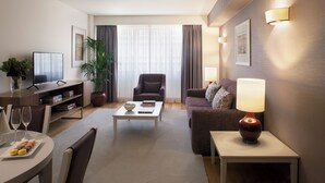 A modern hotel room with a flat-screen TV, a sofa, a dining table with a vase of flowers, and a potted plant.