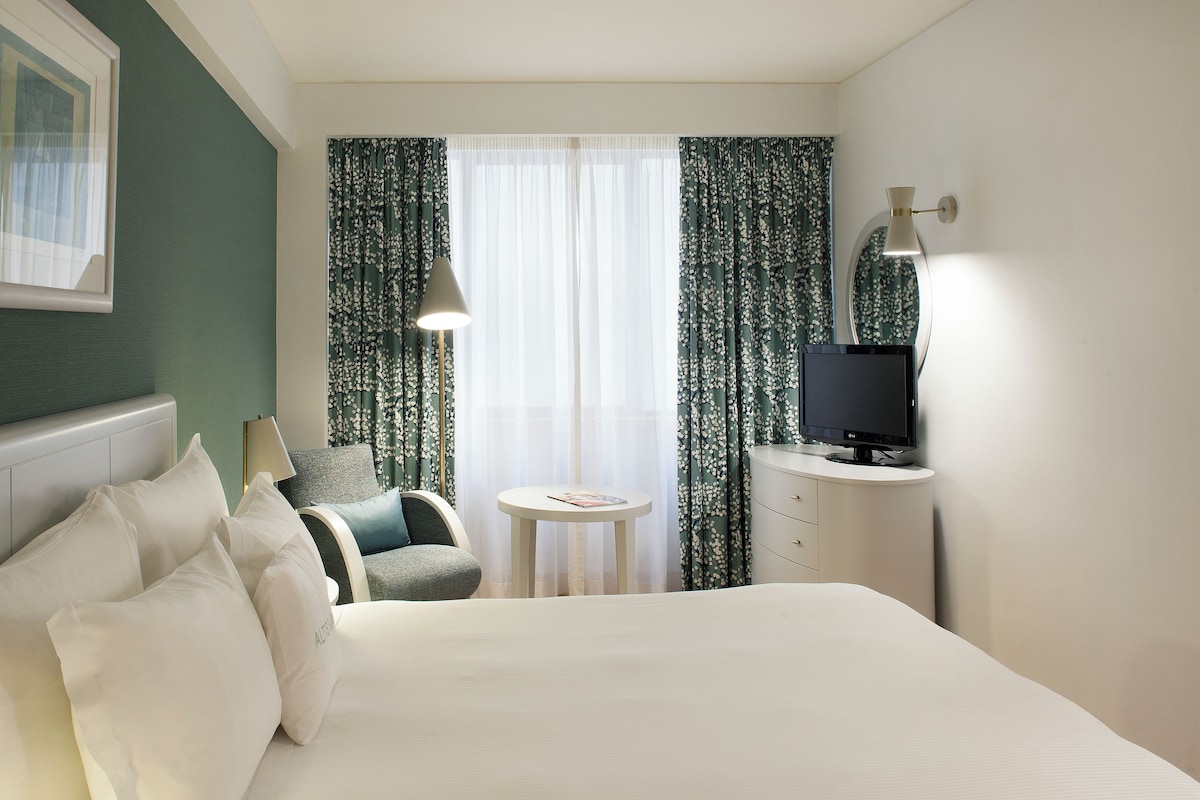 Una habitación de hotel con cama, escritorio con televisor, silla, una mesita y cortinas.