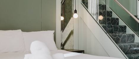 A hotel room with a bed, a rolled towel, a nightstand, a staircase with patterned carpet, and a wall-mounted light fixture.