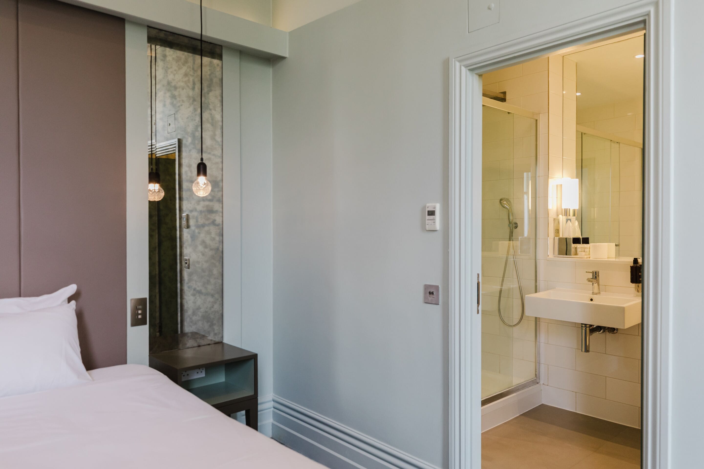 A modern hotel room with a bed, bedside table, and a glass-enclosed shower area.