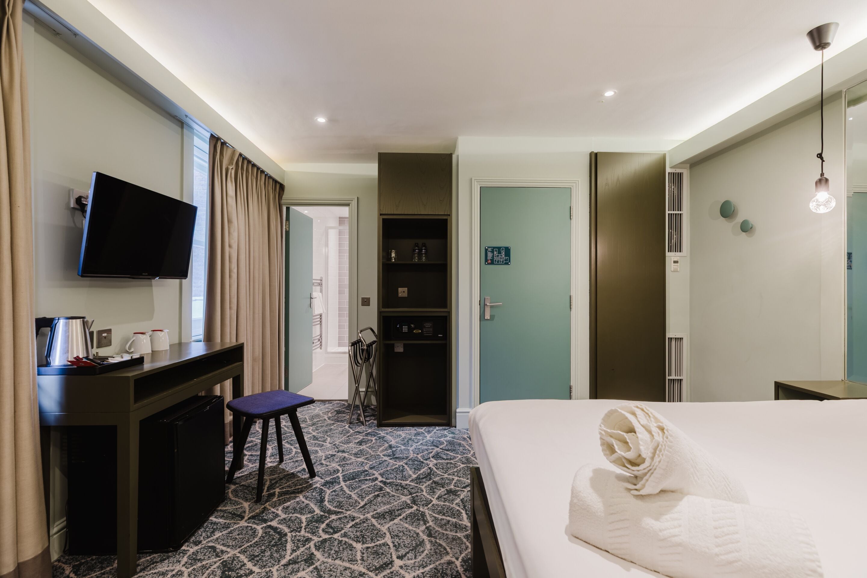 A hotel room with a bed, a desk with a TV, a minibar, and a bathroom door.