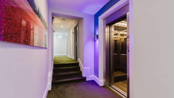 A hallway with a staircase leading to an elevator, a painting on the wall, and a purple-lit wall.