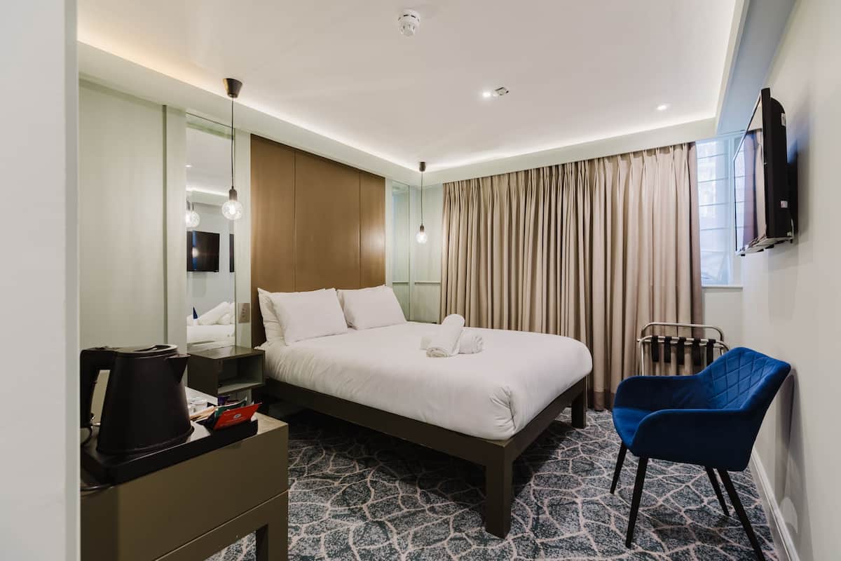 A hotel room with a bed, a desk with a coffee maker, a chair, a TV, and a window with curtains.