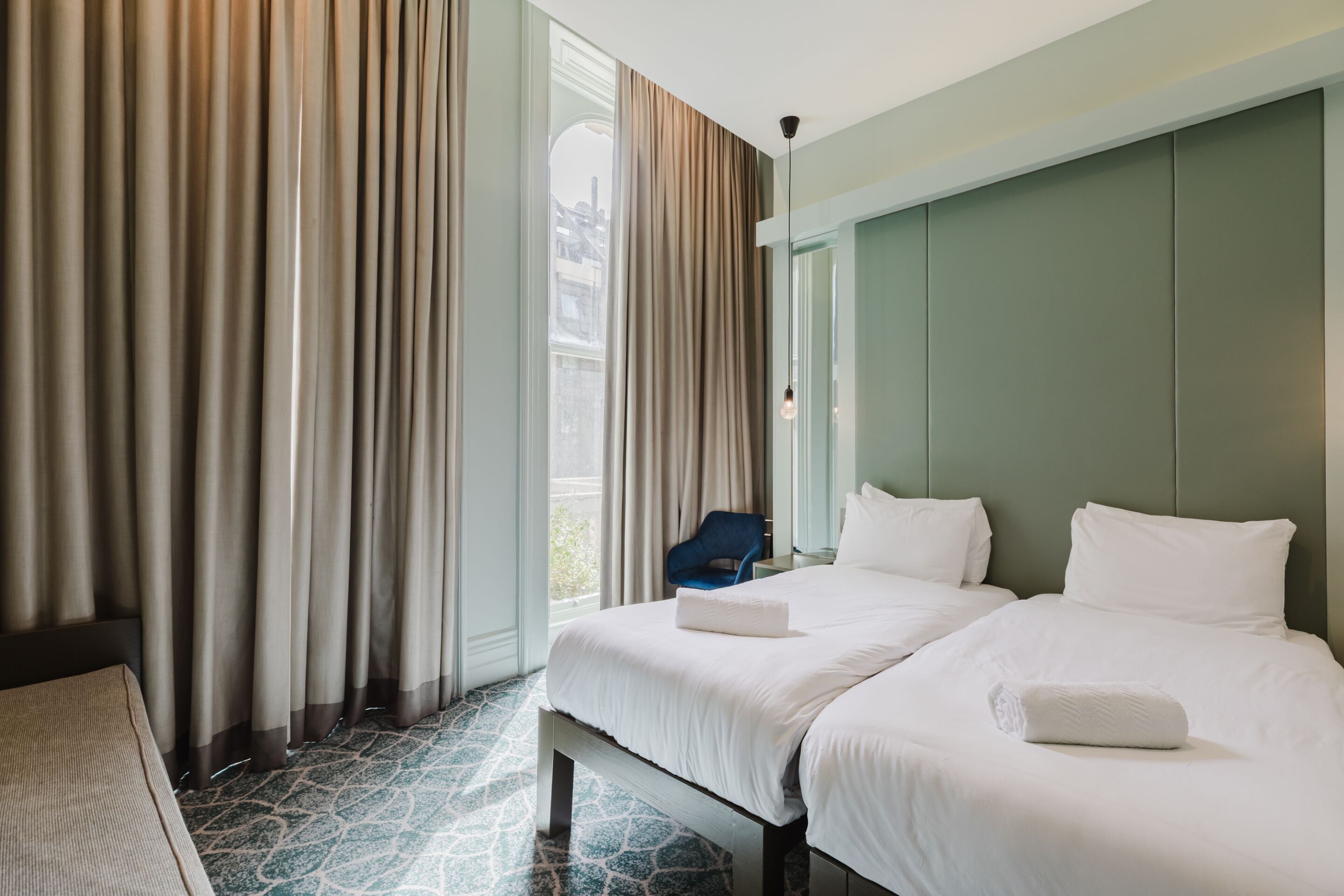 A hotel room with two beds, a large window with curtains, a blue chair, and a patterned floor.