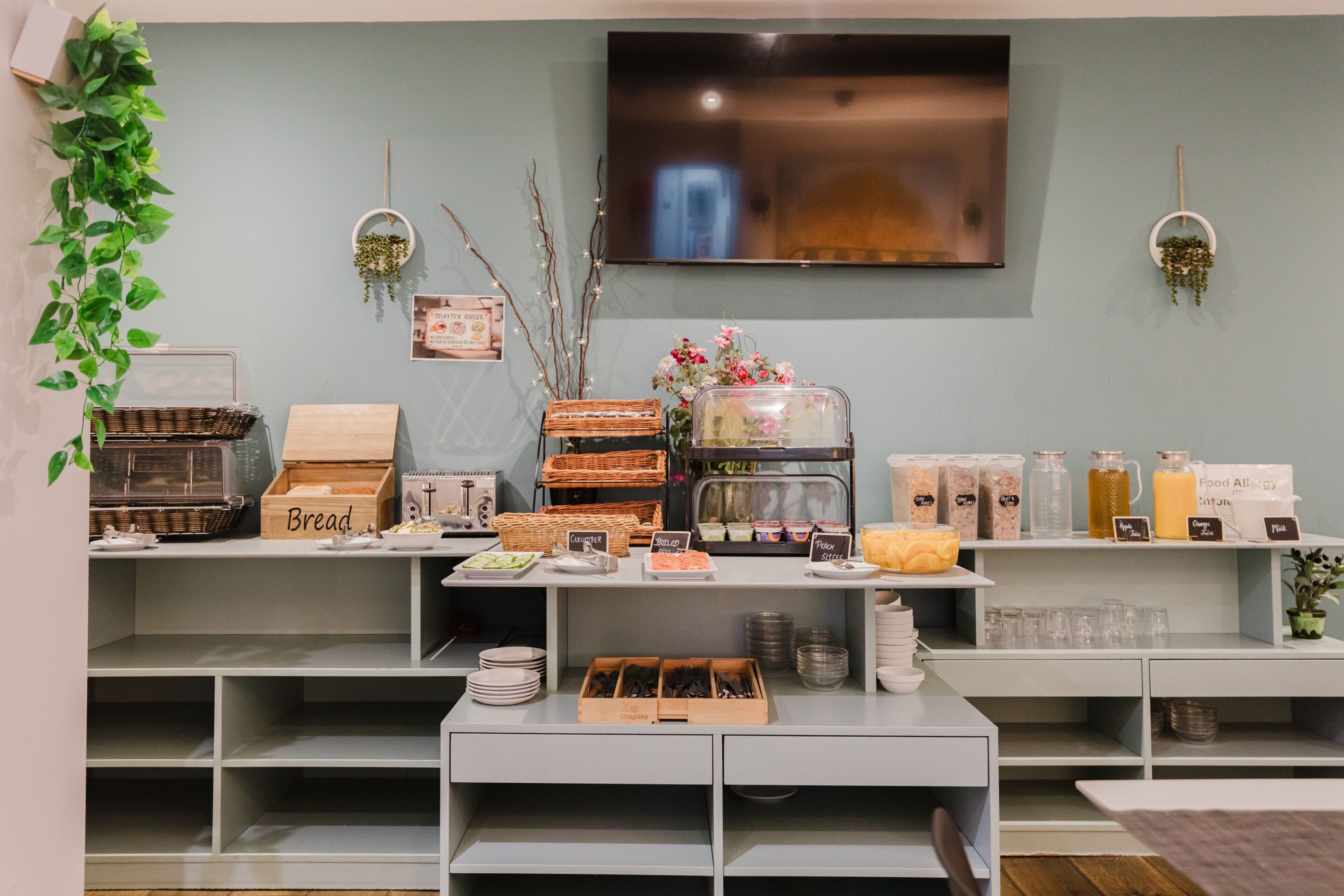 A buffet setup with labeled food stations, a television mounted on the wall, and a potted plant on the left.