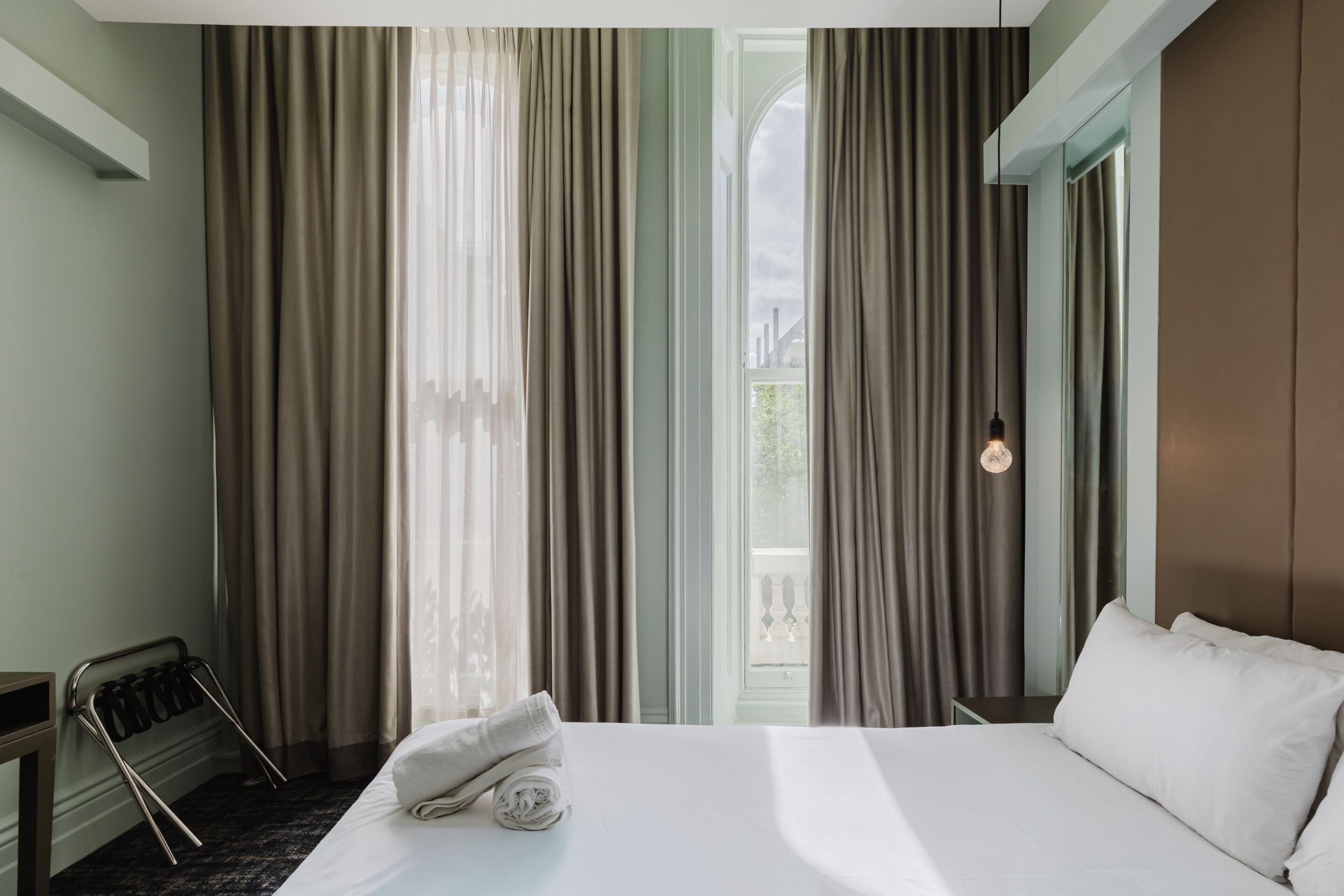 A hotel room with a bed, a window with curtains, a towel on the bed, and a hanging light fixture.