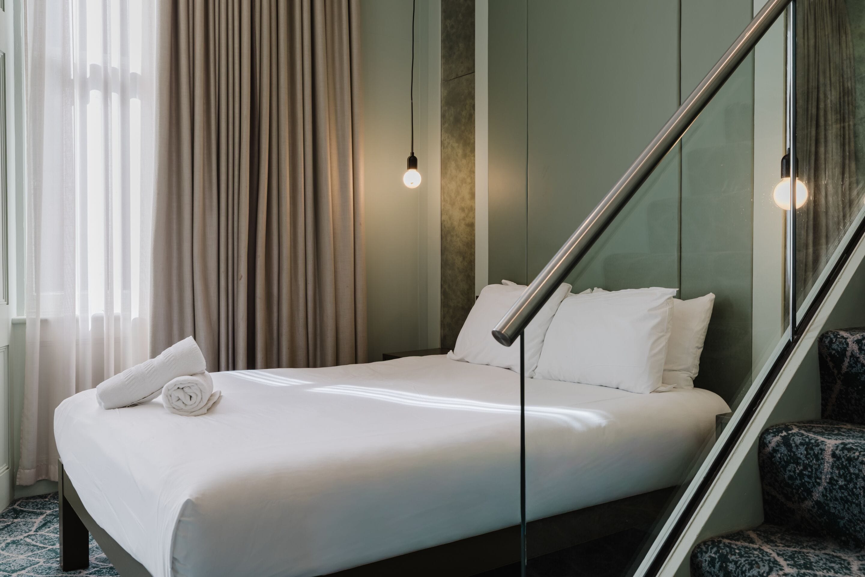 Een hotelkamer met een bed, een stoel, een glazen balustrade en een raam met gordijnen.