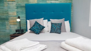 A neatly made bed with a teal headboard, white and teal patterned pillows, and a bedside table with a lamp and remote control. - Paddington Park Hotel (London)