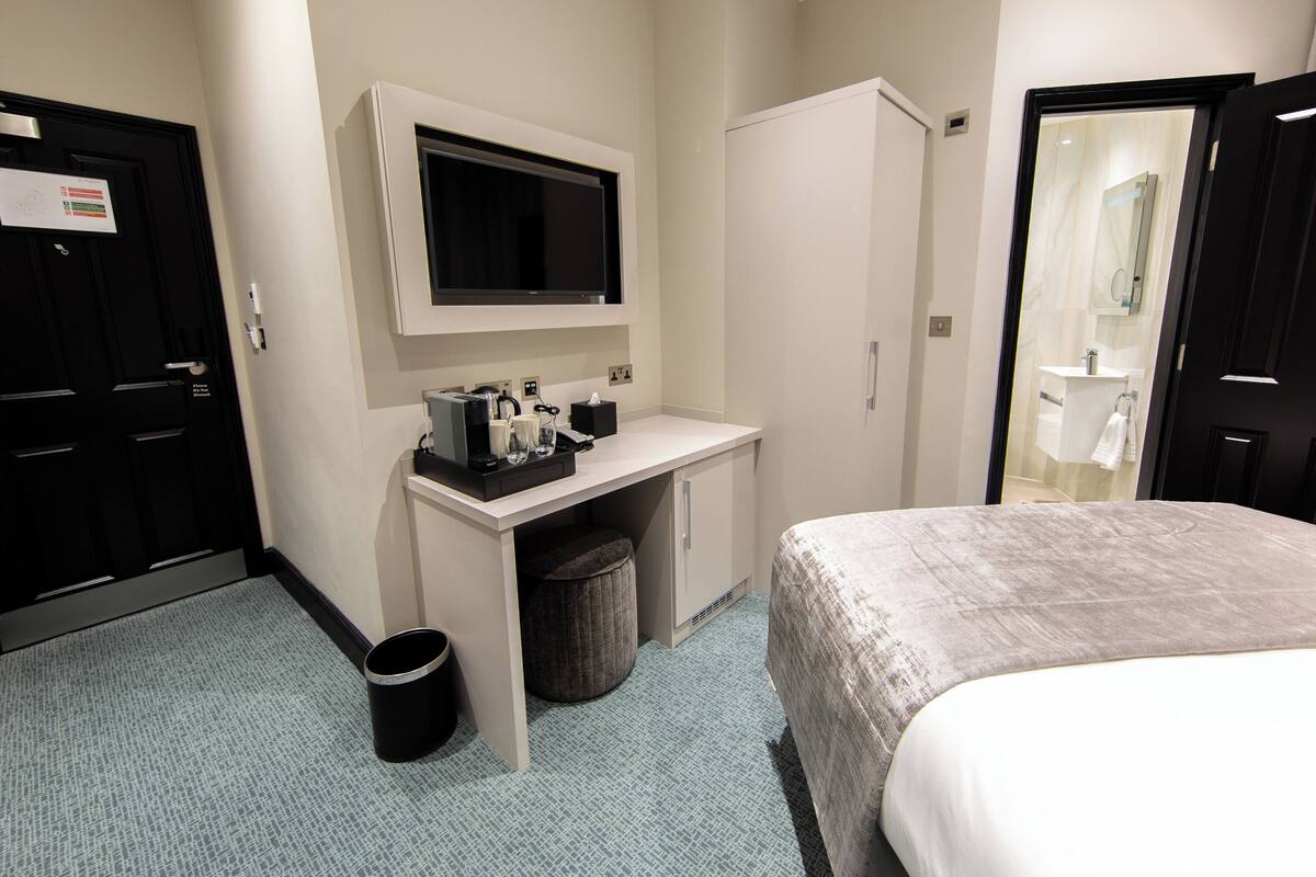 A hotel room with a bed, a desk with a TV, a mirror, and a small trash bin.