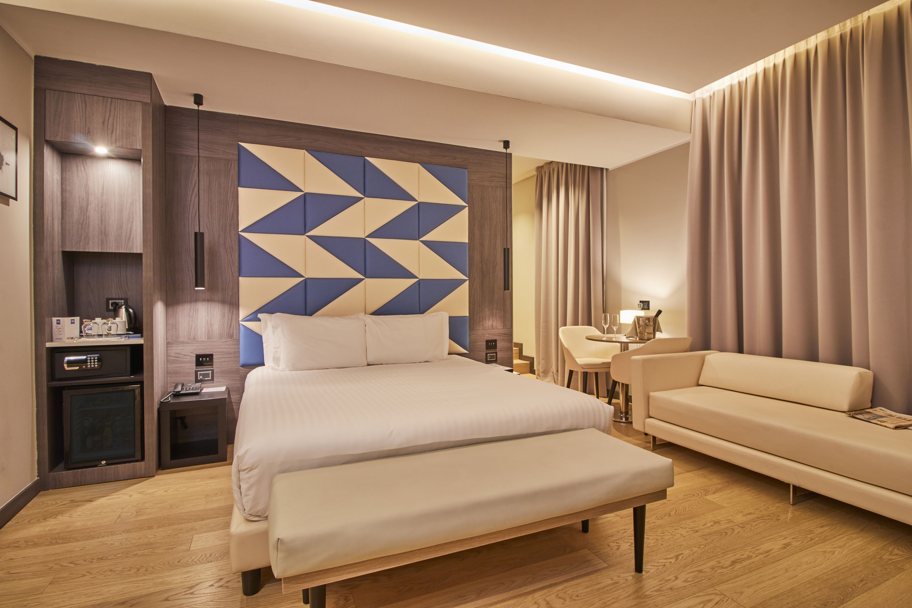 Una moderna camera d'albergo con un letto grande, una poltrona, una scrivania con lampada e un minibar.