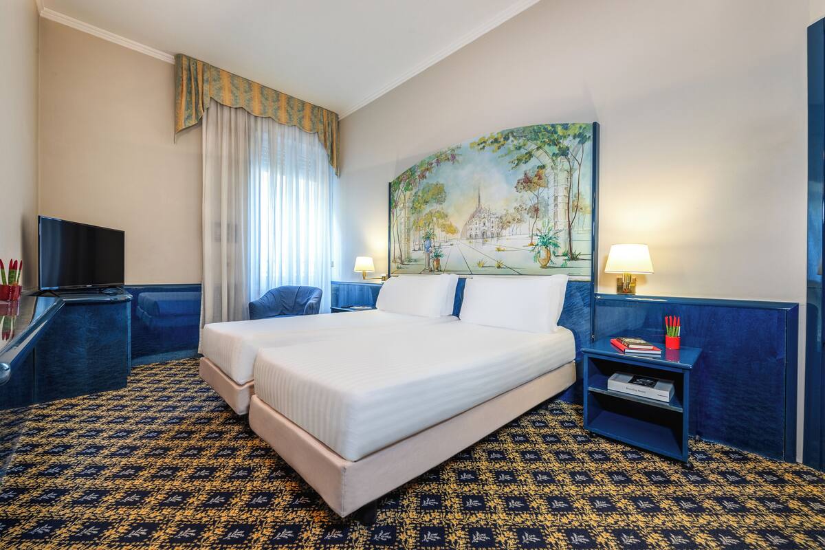 A hotel room with a large bed, two bedside tables, a TV, and a painting on the wall.
