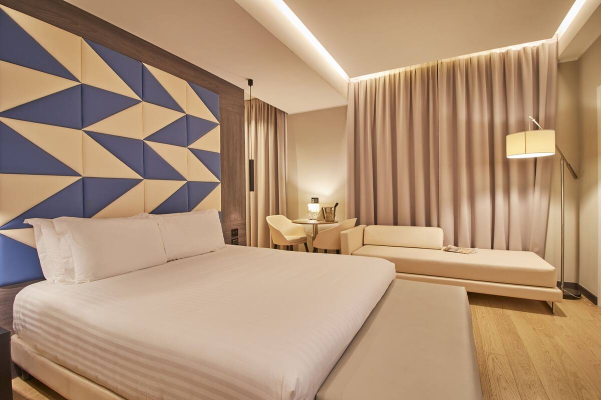 A modern hotel room with a large bed, a sofa, a small table, and a lamp.