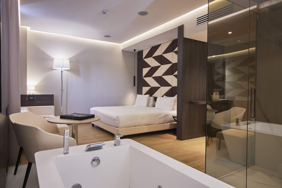 A modern hotel room with a large bed, a glass-enclosed shower, a desk, and a chair.
